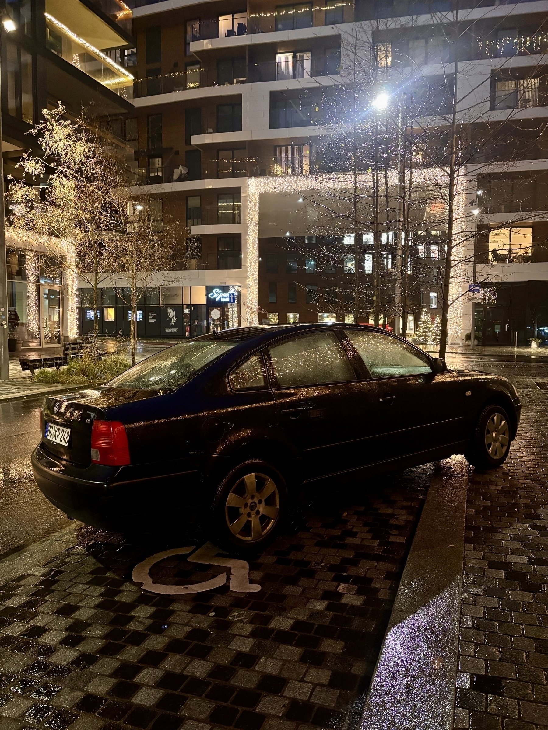 A black car is parked in a handicapped space on a wet street at night, with a building in the background. Front wheel on sidewalk