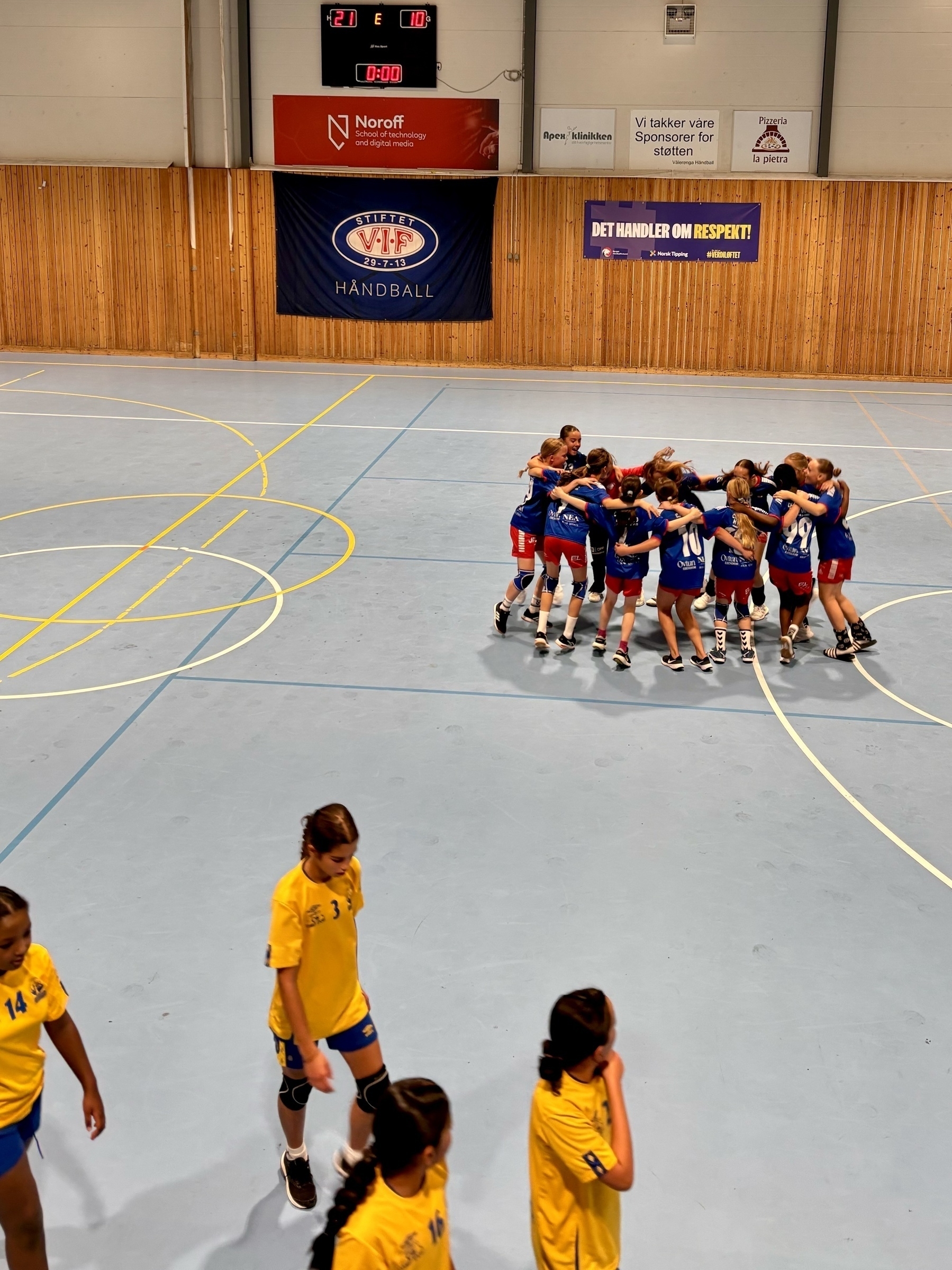 Seierfeiring på håndballbane. Tavla viser 21 - 10 til hjemmelaget (men forskjellen var ikke så stor). 🇬🇧A group of handball players in blue uniforms celebrate together, while players in yellow walk off court. 