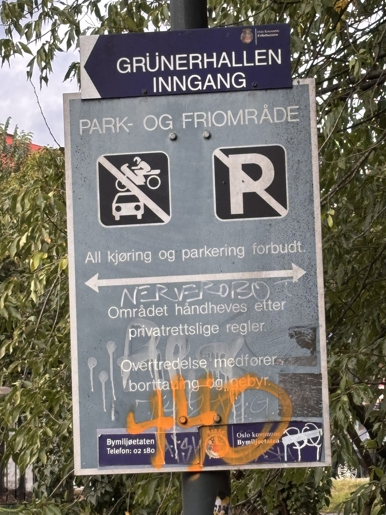 A weathered parking and traffic prohibition sign from Grünerhallen, Oslo, covered in graffiti contrasts with the lush greenery surrounding it.