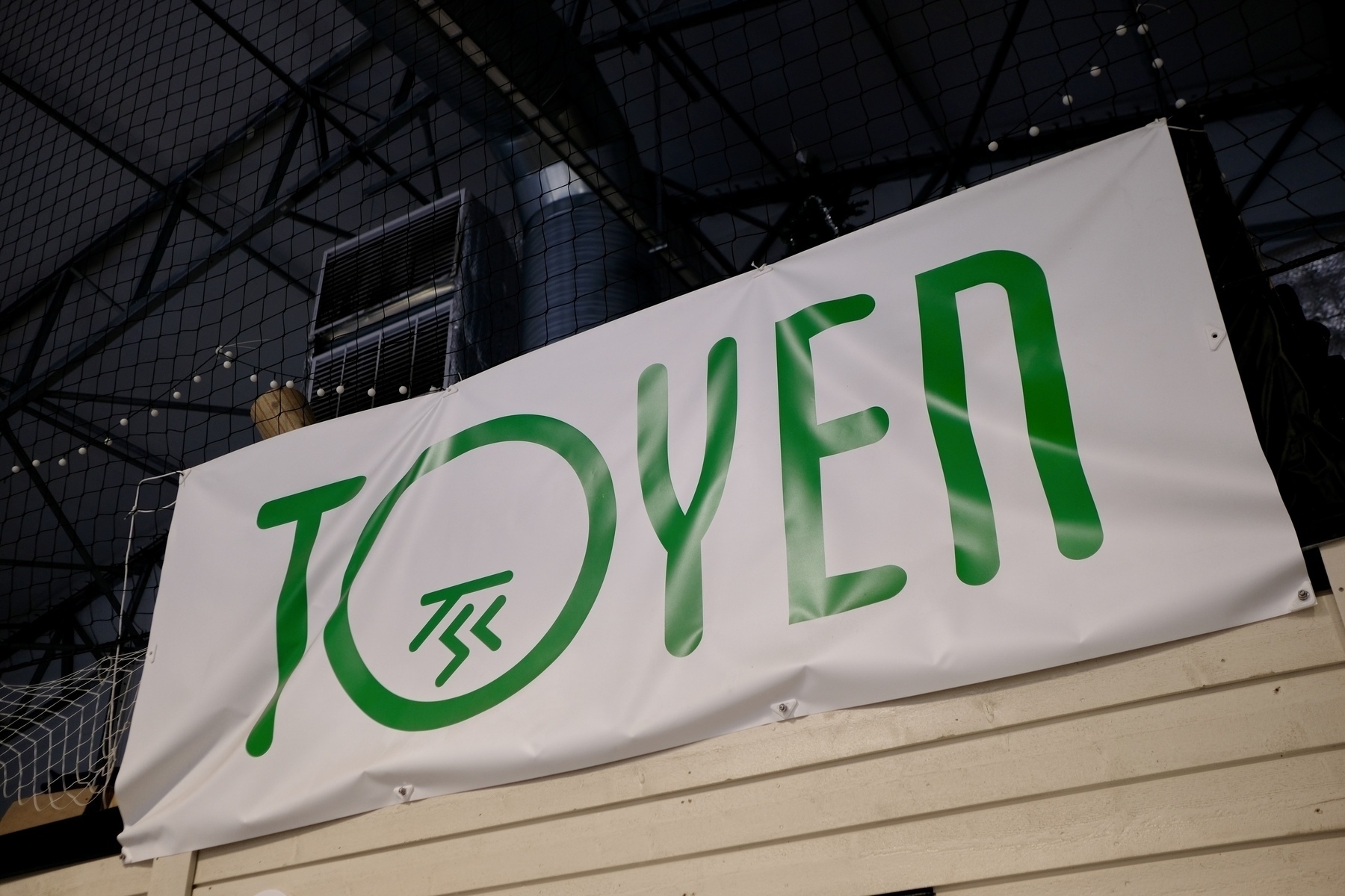 Auto-generated description: A banner with the word TOYEN in green text is displayed against an industrial ceiling backdrop.