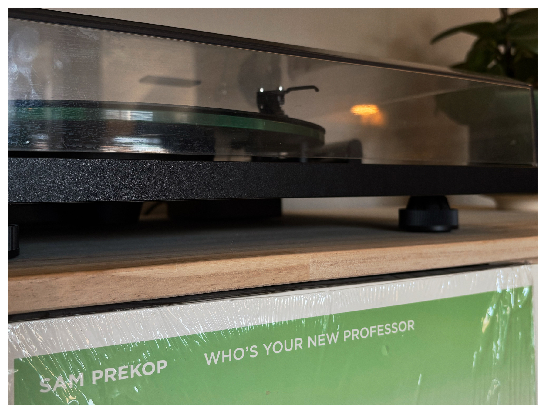 En platespiller med et gjennomsiktig deksel står på en hylle ved siden av et plateomslag med teksten SAM PREKOP WHO'S YOUR NEW PROFESSOR.